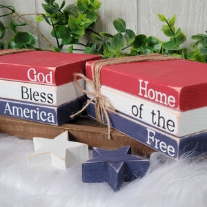 May include: Three stacked wooden blocks painted in red, white, and blue with the words "God Bless America" on one set and "Home of the Free" on the other. The blocks are tied together with twine.