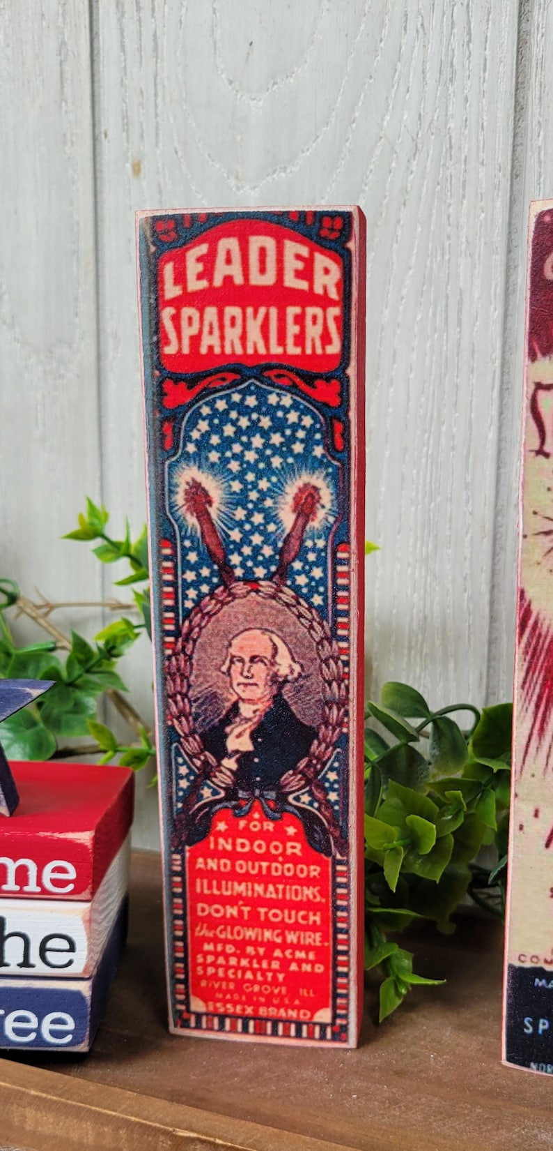 May include: Vintage-style wooden sign with a red, white, and blue design. The sign features a portrait of George Washington and the text "Leader Sparklers" and "For Indoor and Outdoor Illuminations. Don't Touch. The Glowing Wire. Mfd. By Acme Sparkler and Specialty River Grove Ill. Made in USA Essex Brand."