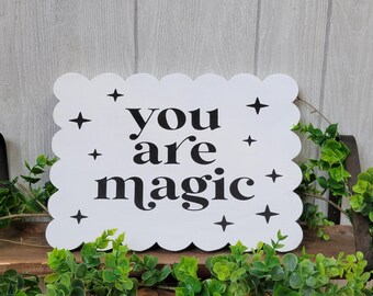 May include: White wooden sign with scalloped edges and black text that reads "you are magic". The sign is decorated with small black stars.