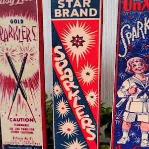 May include: Three vintage wooden boxes with red, white, and blue labels. The labels feature the words "Sparkler", "Gold Parklers", and "Star Brand". The boxes are decorated with images of fireworks and stars.