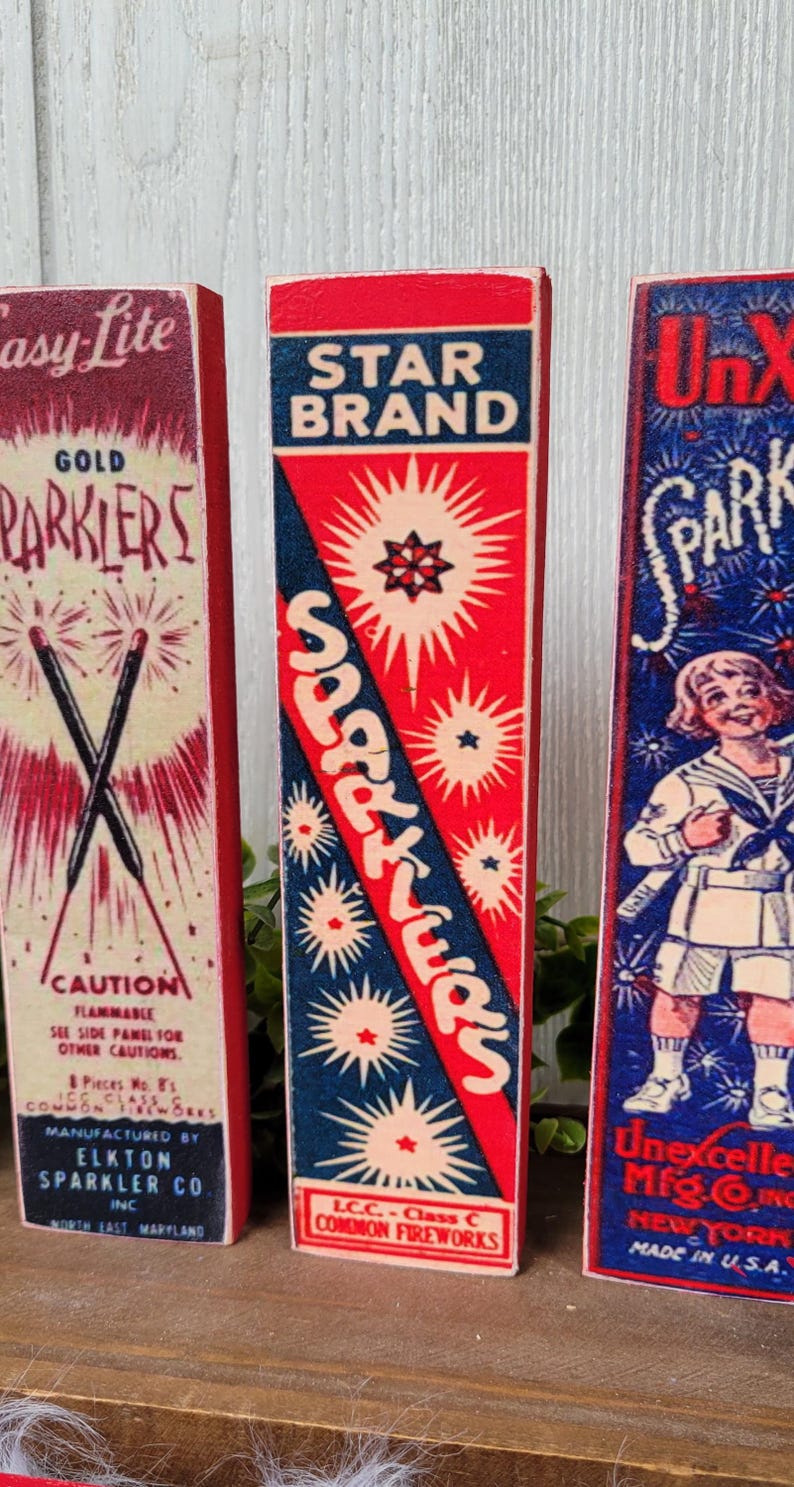 May include: Three vintage wooden boxes with red, white, and blue labels. The labels feature the words "Sparkler", "Gold Parklers", and "Star Brand". The boxes are decorated with images of fireworks and stars.