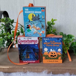 May include: Three miniature book ornaments with orange ribbon hangers. The books feature covers with the titles "The Haunted House", "Goosebumps", and "It's the Great Pumpkin, Charlie Brown". The ornaments are likely for Halloween or book lovers.