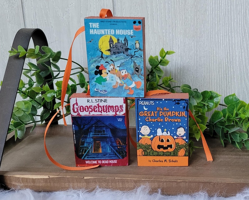 May include: Three miniature book ornaments with orange ribbon hangers. The books feature covers with the titles "The Haunted House", "Goosebumps", and "It's the Great Pumpkin, Charlie Brown". The ornaments are likely for Halloween or book lovers.