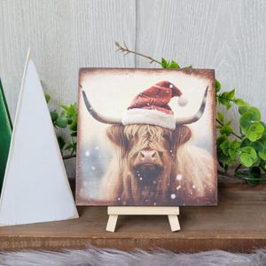 May include: A square wooden sign with a painting of a Highland cow wearing a Santa hat. The cow is brown and white with a long, shaggy coat. The background is a snowy landscape. The sign is on a small wooden easel.