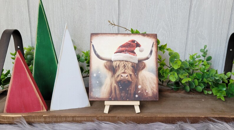 May include: A square wooden sign with a painting of a Highland cow wearing a Santa hat. The cow is brown and white with a long, shaggy coat. The background is a snowy landscape. The sign is on a small wooden easel.