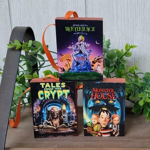 May include: Three small, rectangular wooden ornaments featuring movie posters: "Beetlejuice," "Tales from the Crypt," and "Monster House." Each ornament has an orange ribbon for hanging. The "Beetlejuice" ornament shows a man in a striped suit.