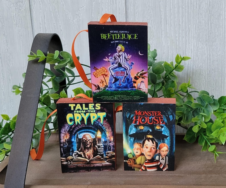May include: Three small, rectangular wooden ornaments featuring movie posters: "Beetlejuice," "Tales from the Crypt," and "Monster House." Each ornament has an orange ribbon for hanging. The "Beetlejuice" ornament shows a man in a striped suit.