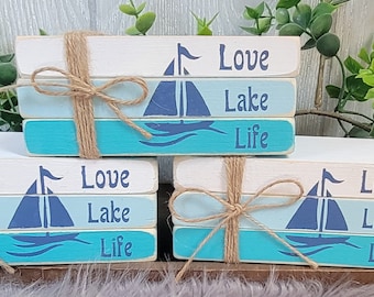 Puede incluir: Tres bloques de madera apilados con un acabado de pintura degradado azul, blanco y azul claro. Cada bloque tiene un gráfico de velero y las palabras "Love Lake Life" escritas en negro.