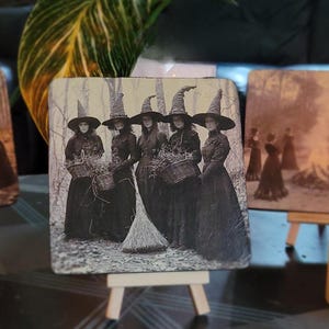 Puede incluir: Posavasos cuadrado con una imagen vintage en blanco y negro de cinco mujeres con sombreros de bruja y vestidos largos, sosteniendo cestas y una escoba. La imagen est&aacute; ambientada en un fondo de bosque, evocando un tema de Halloween o g&oacute;tico.