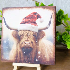 May include: A Highland cow wearing a red and white plaid Santa hat with a white fur trim. The cow is standing in a snowy field.
