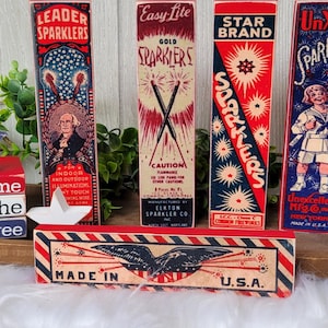 May include: A wooden sign with red, white, and blue stripes reads "Home of the Free".  The sign is decorated with a red, white, and blue star and is sitting on a wooden shelf with five vintage boxes of sparklers. The boxes are labeled "Leader Sparkler", "Easy Lite Gold Sparkler", "Star Brand Sparkler", "UnXid Sparkler", and "Made in U.S.A.".