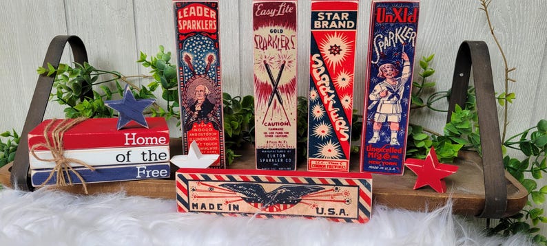 May include: A wooden sign with red, white, and blue stripes reads "Home of the Free".  The sign is decorated with a red, white, and blue star and is sitting on a wooden shelf with five vintage boxes of sparklers. The boxes are labeled "Leader Sparkler", "Easy Lite Gold Sparkler", "Star Brand Sparkler", "UnXid Sparkler", and "Made in U.S.A.".