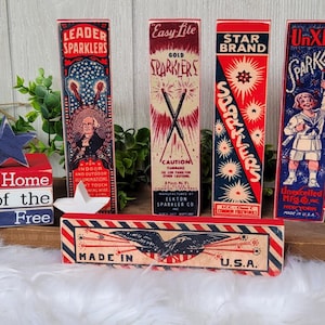 May include: A set of five vintage-style wooden blocks with red, white, and blue patriotic designs. The blocks feature images of sparklers, fireworks, and a child holding a sparkler. The blocks are stacked on a wooden surface and have the text "Home of the Free" and "Made in U.S.A." printed on them.