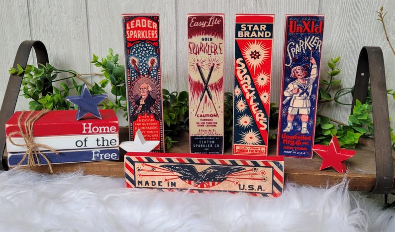 May include: A set of five vintage-style wooden blocks with red, white, and blue patriotic designs. The blocks feature images of sparklers, fireworks, and a child holding a sparkler. The blocks are stacked on a wooden surface and have the text "Home of the Free" and "Made in U.S.A." printed on them.