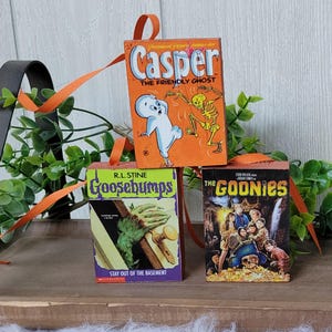 May include: Three miniature book ornaments featuring the covers of "Casper the Friendly Ghost," "Goosebumps," and "The Goonies." Each ornament has an orange ribbon for hanging. The "Casper" ornament is orange, the "Goosebumps" ornament is green, and "The Goonies" is dark.