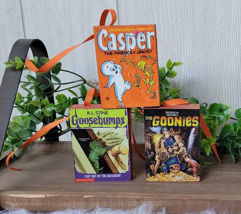 May include: Three miniature book ornaments featuring the covers of "Casper the Friendly Ghost," "Goosebumps," and "The Goonies." Each ornament has an orange ribbon for hanging. The "Casper" ornament is orange, the "Goosebumps" ornament is green, and "The Goonies" is dark.