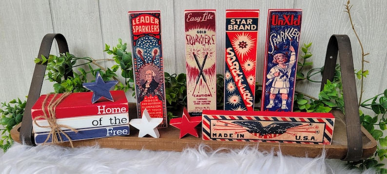 May include: A wooden tray with red, white, and blue decorations, including a sign that reads "Home of the Free", and five vintage-style sparkler boxes with red, white, and blue designs. The boxes have text that reads "Leader Sparkler", "Easy Lite Gold Sparkler", "Star Brand Sparkler", "UnXId Sparkler", and "Made in U.S.A."