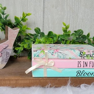 May include: A stack of three decorative books with a floral design. The top book has the text "Spring is in full bloom" on the front. The books are tied together with a white ribbon.