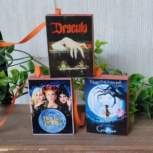 May include: Three Halloween-themed wooden book ornaments with orange ribbon hangers. The books feature the covers of "Dracula", "Hocus Pocus", and "Coraline". The "Hocus Pocus" book has a blue circle with the movie title.