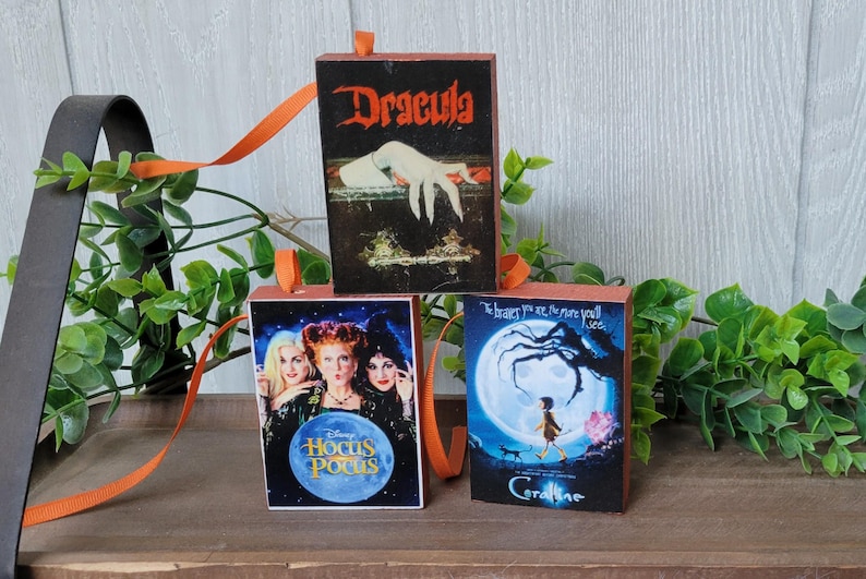 May include: Three Halloween-themed wooden book ornaments with orange ribbon hangers. The books feature the covers of "Dracula", "Hocus Pocus", and "Coraline". The "Hocus Pocus" book has a blue circle with the movie title.