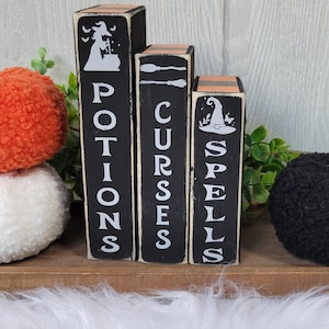 May include: Halloween-themed wooden book set with the words "Potions", "Curses", and "Spells" in white lettering. Each book has a different witchy graphic. Decorative orange and black yarn pumpkins sit on either side of the books.