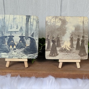 Puede incluir: Dos placas cuadradas de madera sobre peque&ntilde;os caballetes. Una representa un grupo de brujas con capas y sombreros alrededor de velas. La otra muestra a mujeres con vestidos largos alrededor de una hoguera. Im&aacute;genes de estilo vintage.