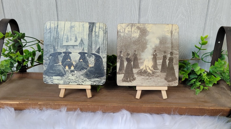 Puede incluir: Dos placas cuadradas de madera sobre peque&ntilde;os caballetes. Una representa un grupo de brujas con capas y sombreros alrededor de velas. La otra muestra a mujeres con vestidos largos alrededor de una hoguera. Im&aacute;genes de estilo vintage.