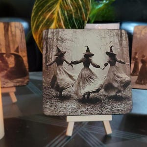 Puede incluir: Posavasos cuadrado con una imagen en tonos sepia de tres brujas bailando en un bosque, tomadas de la mano. Las brujas visten vestidos largos y sombreros puntiagudos. El posavasos se exhibe en un peque&ntilde;o caballete de madera, con otros posavasos similares en el fondo.