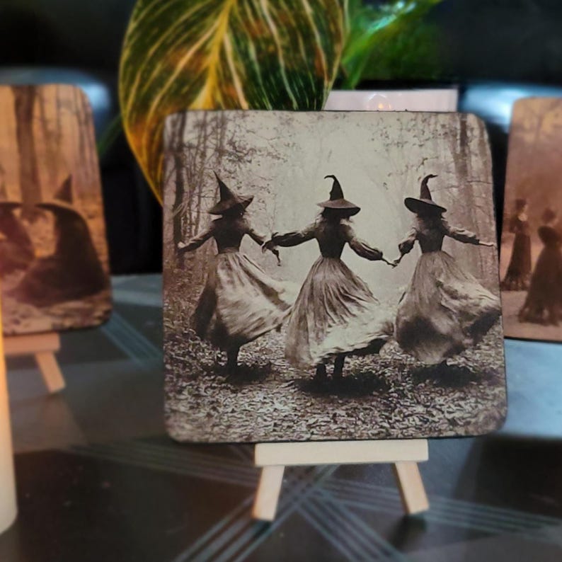 Puede incluir: Posavasos cuadrado con una imagen en tonos sepia de tres brujas bailando en un bosque, tomadas de la mano. Las brujas visten vestidos largos y sombreros puntiagudos. El posavasos se exhibe en un peque&ntilde;o caballete de madera, con otros posavasos similares en el fondo.