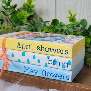 May include: A stack of three wooden books with a spring theme. The top book is yellow and says "April showers bring May flowers". The middle book is white with blue raindrops and the bottom book is blue with white text.