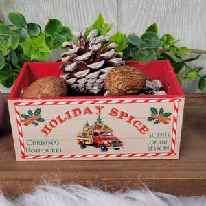 May include: A red and cream-colored wooden box with the words "Holiday Spice" and "Christmas Potpourri" printed on the front. The box contains a pine cone, nuts, and decorative accents. Silver glitter ornaments and greenery are in the background.