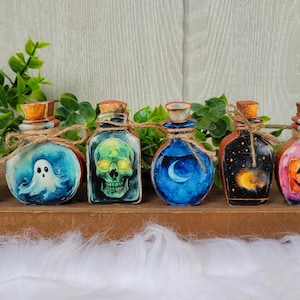 May include: A collection of eight decorative glass bottles with cork stoppers, each featuring a unique Halloween-themed design. The bottles are tied with twine and showcase images of pumpkins, candy corn, ghosts, skulls, and jack-o'-lanterns.