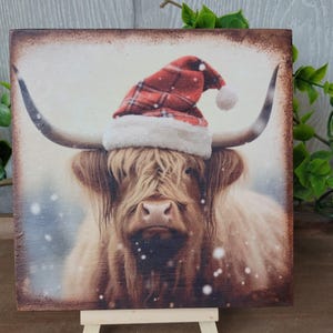 May include: A Highland cow wearing a red and white Santa hat with a white pom-pom on a snowy background.