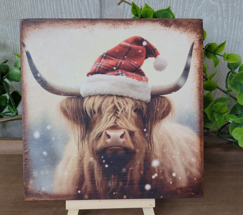 May include: A Highland cow wearing a red and white Santa hat with a white pom-pom on a snowy background.