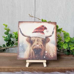 May include: A square wooden sign with a painting of a Highland cow wearing a red and white Santa hat. The cow is standing in a snowy field. The sign is on a small wooden easel.