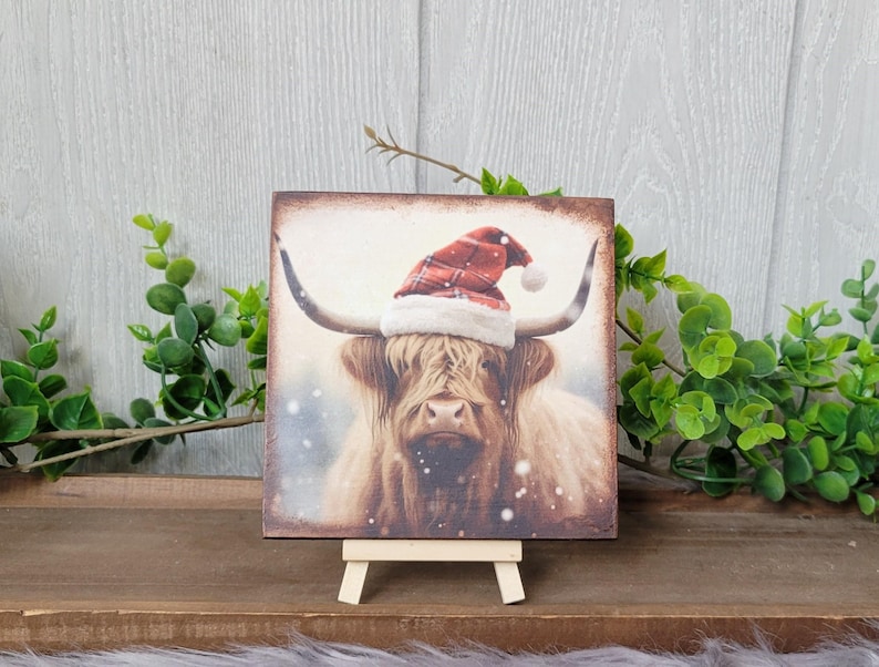 May include: A square wooden sign with a painting of a Highland cow wearing a red and white Santa hat. The cow is standing in a snowy field. The sign is on a small wooden easel.