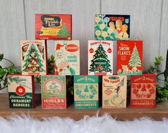 May include: A collection of vintage Christmas ornament boxes in various colors, including red, green, and teal. The boxes feature illustrations of Christmas trees, ornaments, and Santa Claus. Some boxes have text like "Shiny Brite" and "Snowflakes".