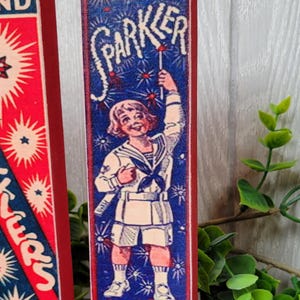 May include: A vintage wooden sign with a red border and blue background featuring a child holding a sparkler. The sign reads "UnXld SPARKLER Unexcelled Mfg. Co. Inc. NEW YORK MADE IN U.S.A."