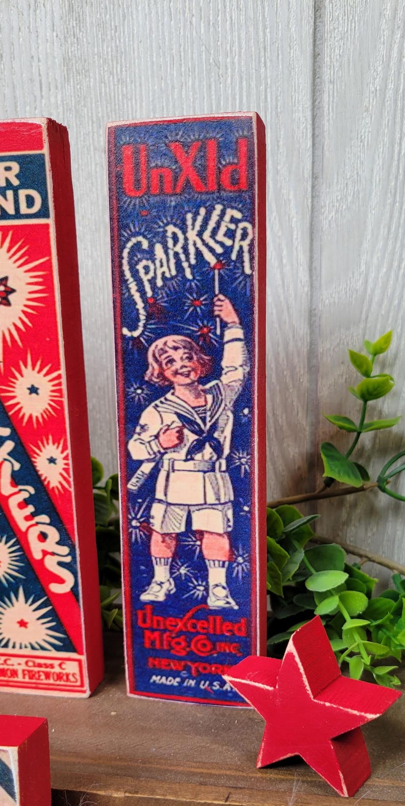 May include: A vintage wooden sign with a red border and blue background featuring a child holding a sparkler. The sign reads "UnXld SPARKLER Unexcelled Mfg. Co. Inc. NEW YORK MADE IN U.S.A."