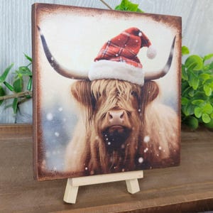 May include: A brown and white Highland cow wearing a red and white plaid Santa hat. The cow is standing in a snowy field. The image is printed on a wooden plaque with a small easel.