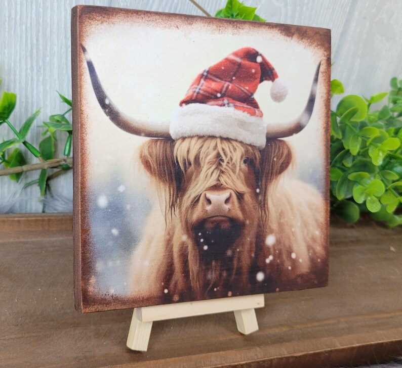 May include: A brown and white Highland cow wearing a red and white plaid Santa hat. The cow is standing in a snowy field. The image is printed on a wooden plaque with a small easel.