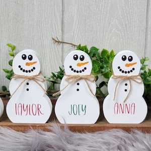 May include: Five white wooden snowman ornaments with names written on the bottom. The names are Josh, Taylor, Joly, Anna, and Jonathan.