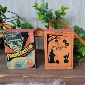 May include: Two Halloween-themed ornaments shaped like vintage books. One features Frankenstein's monster, the other a witch and pumpkins. Both have orange ribbon handles and are set against a backdrop of greenery.