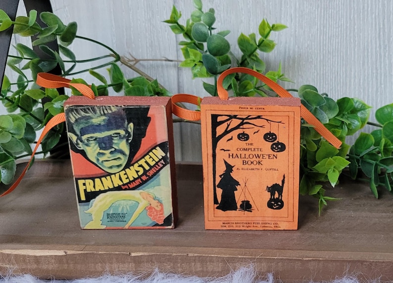 May include: Two Halloween-themed ornaments shaped like vintage books. One features Frankenstein's monster, the other a witch and pumpkins. Both have orange ribbon handles and are set against a backdrop of greenery.