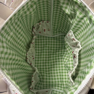 May include: The interior of a tote bag featuring a green and white gingham check pattern lining. The bag includes multiple interior pockets with elasticated edges, decorated with a white fabric print featuring small red cherry illustrations.
