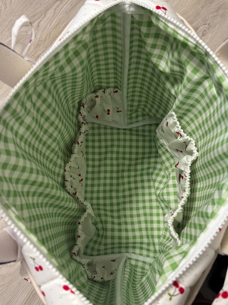 May include: The interior of a tote bag featuring a green and white gingham check pattern lining. The bag includes multiple interior pockets with elasticated edges, decorated with a white fabric print featuring small red cherry illustrations.