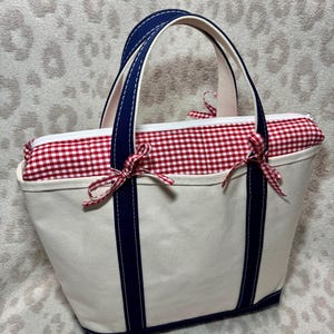 May include: A cream-coloured canvas tote bag featuring two dark navy blue vertical stripes and matching handles. The top opening is secured with a white zip and a red and white gingham fabric insert, accented by two small red and white gingham bows.