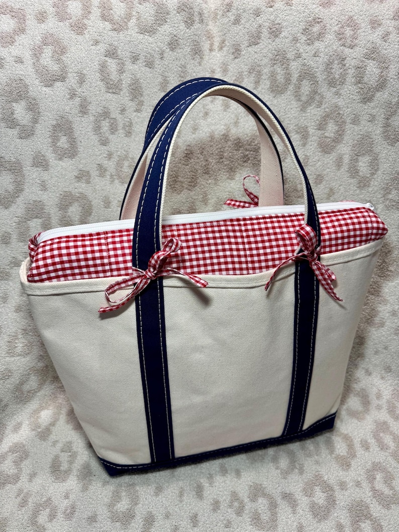 May include: A cream-coloured canvas tote bag featuring two dark navy blue vertical stripes and matching handles. The top opening is secured with a white zip and a red and white gingham fabric insert, accented by two small red and white gingham bows.