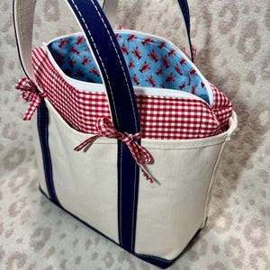 May include: A cream-coloured canvas tote bag featuring navy blue vertical stripes and handles. The top section has a red and white gingham fabric border with small tied bows on the sides. The interior lining is light blue with a red lobster print.