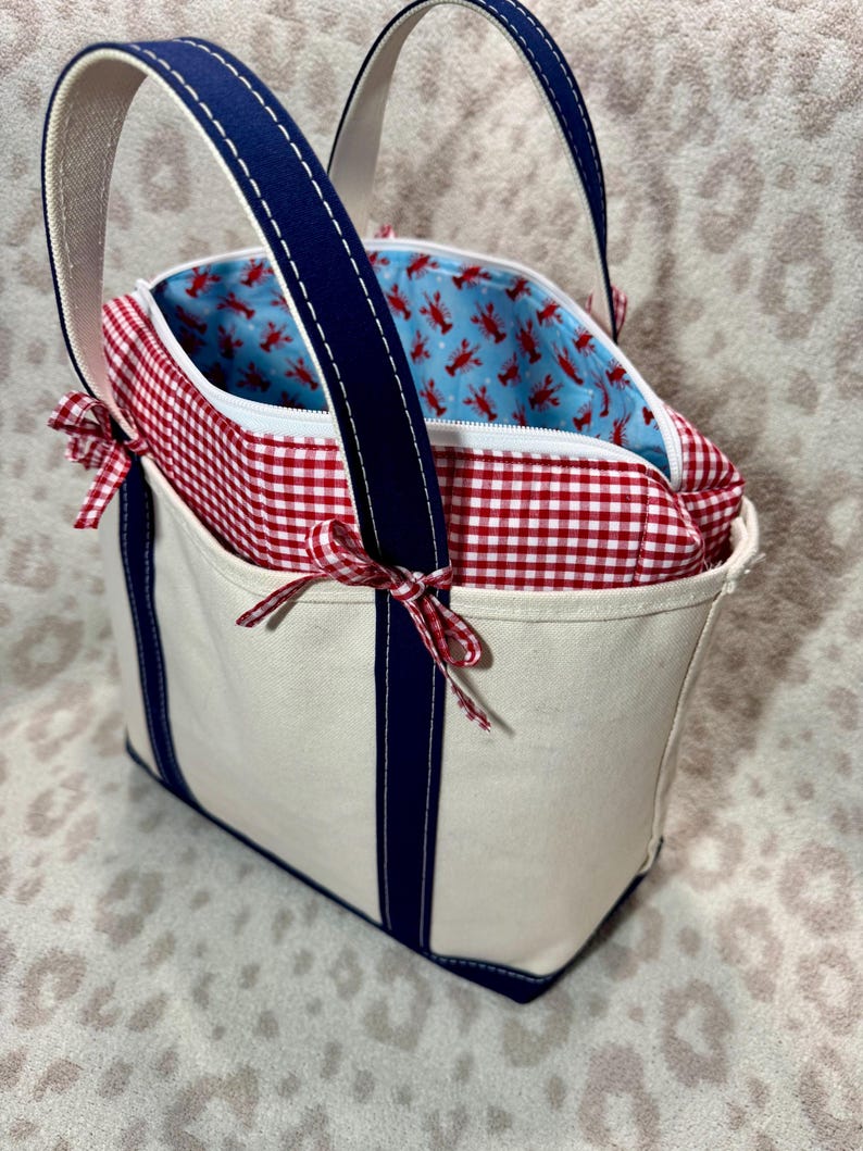 May include: A cream-coloured canvas tote bag featuring navy blue vertical stripes and handles. The top section has a red and white gingham fabric border with small tied bows on the sides. The interior lining is light blue with a red lobster print.
